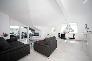 A modern living room with a sofa, a dining area with chairs, a TV, and a view of the outdoors. - Holiday apartment with 2 bedrooms (Oberstaufen)
