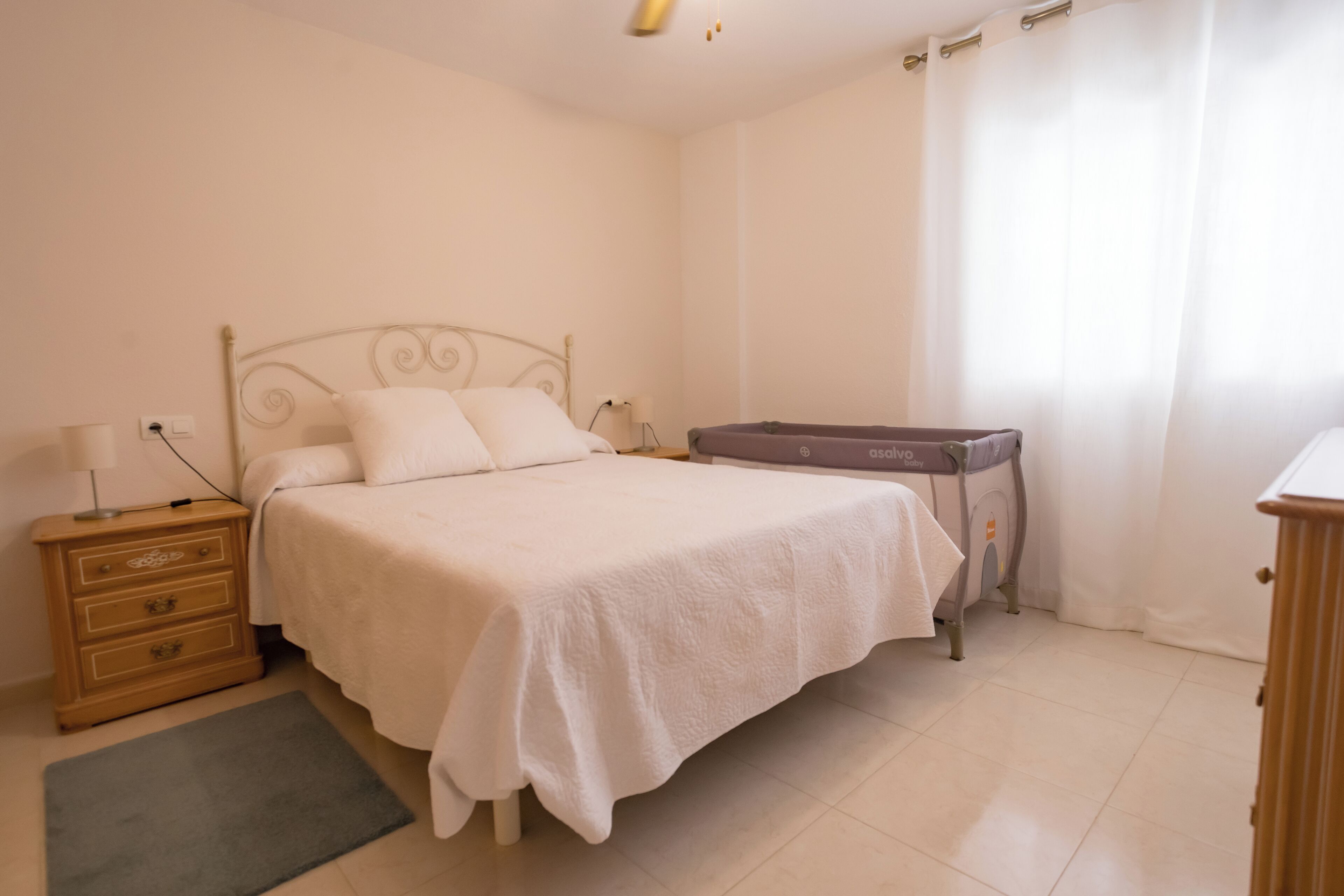 A bedroom with a bed, a nightstand, a dresser, a baby bassinet, and a window with curtains.