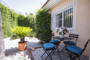 An outdoor seating area with a table and chairs, a potted plant, and a window with white bars.