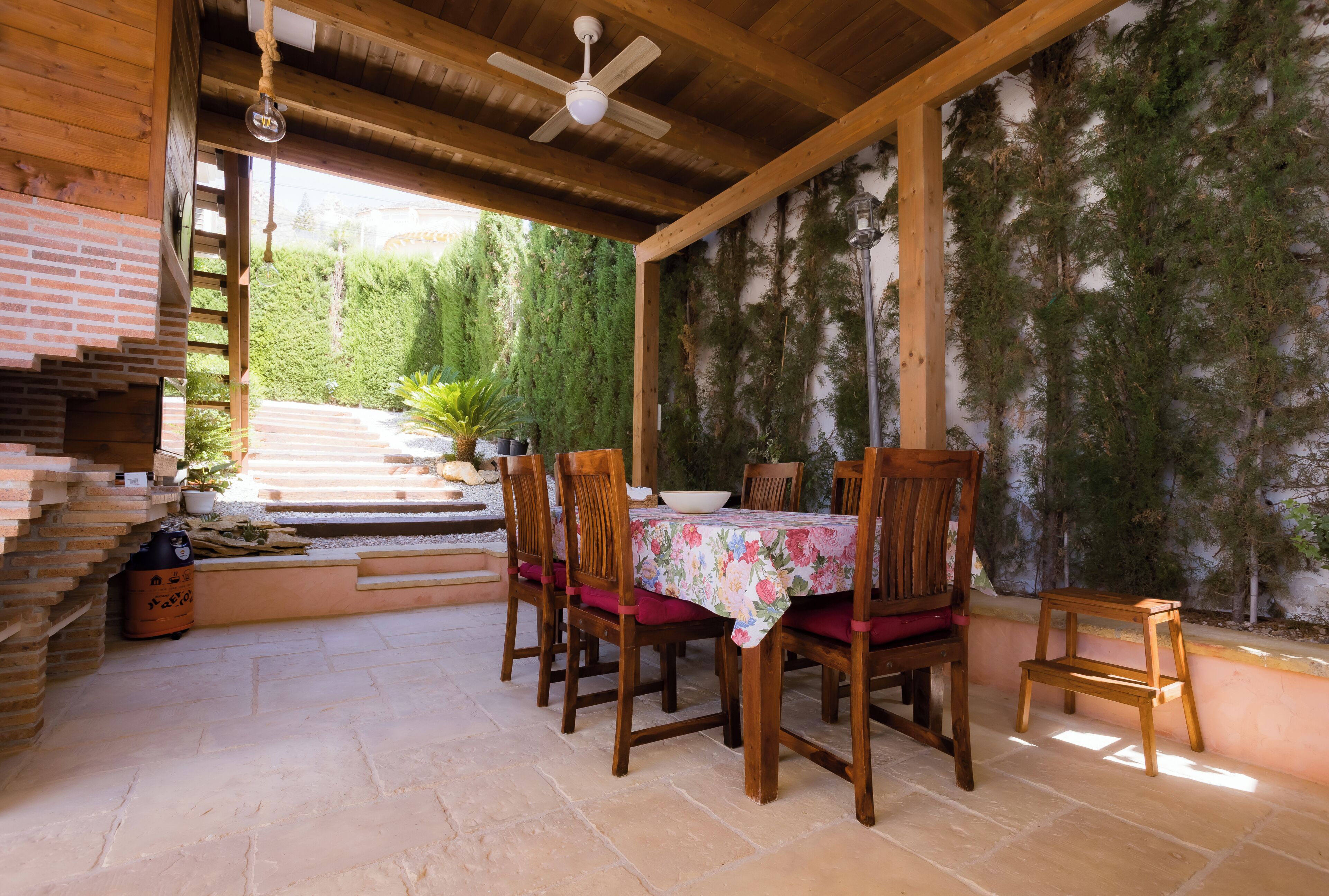 Un espace extérieur abrité, aménagé pour les repas, avec une table et des chaises, un ventilateur de plafond et une vue sur un jardin avec des marches descendant vers le bas.