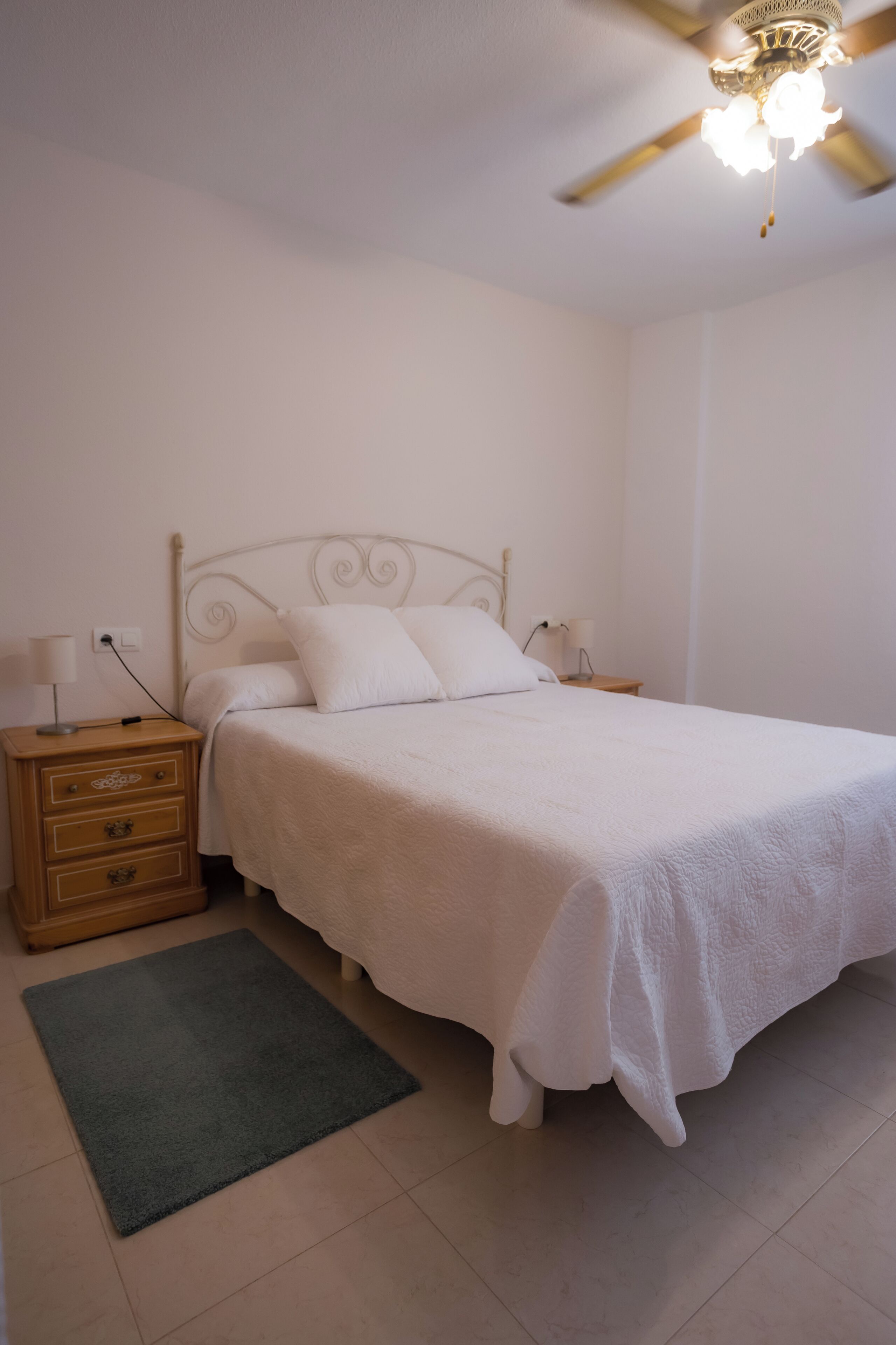 Une chambre à coucher comprenant un lit, une table de chevet, une commode, un ventilateur de plafond et un tapis.