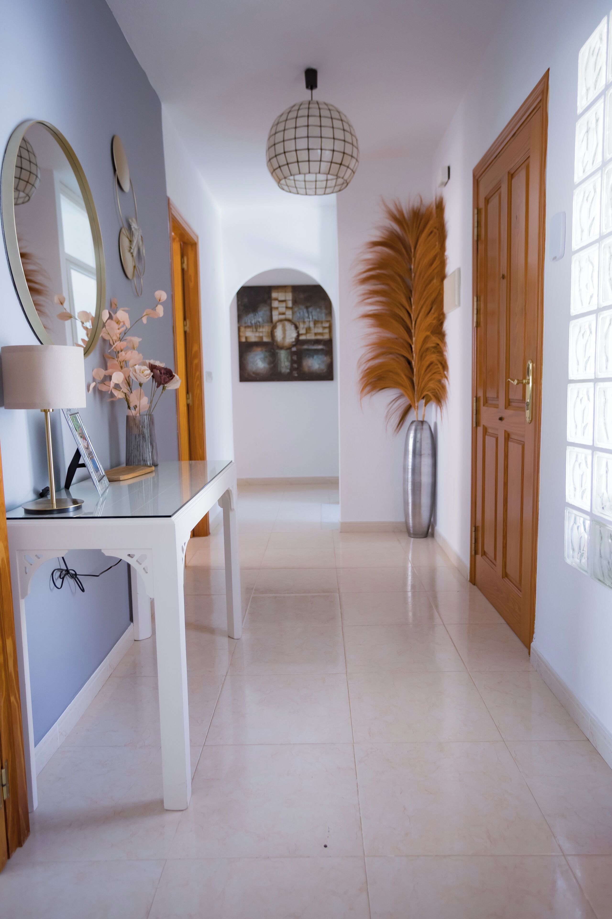 A hallway with a white floor, a table with a lamp, a mirror, and a vase with dried flowers.