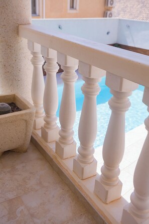 A balcony with a white balustrade, a potted plant, and a glimpse of a pool.