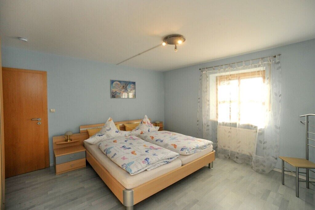 Un dormitorio con cama doble, armario de madera, mesita de noche pequeña, una silla, ventana con cortinas e iluminación en el techo.