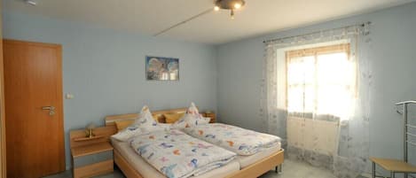 A bedroom with a double bed, a wooden wardrobe, a small bedside table, a chair, a window with curtains, and a ceiling light fixture.