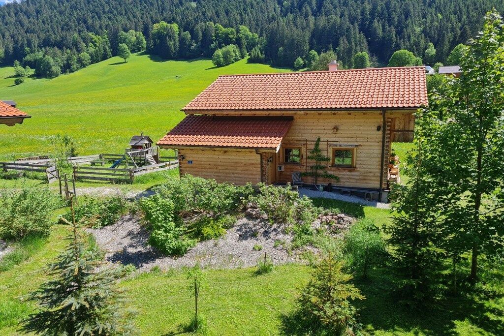 A wooden cabin with a red-tiled roof, surrounded by lush green fields and forested hills.