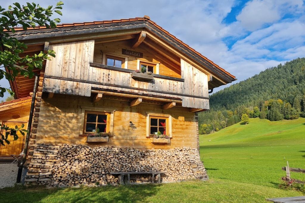 A wooden chalet with a balcony, a stone foundation, and a view of a green valley.
