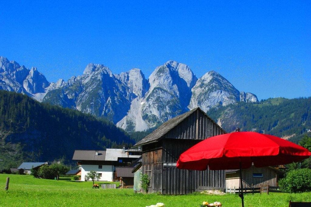 Ländliche Szenerie mit einem hölzernen Bauernhaus und einem roten Regenschirm, umgeben von grünen Feldern und Bergen.