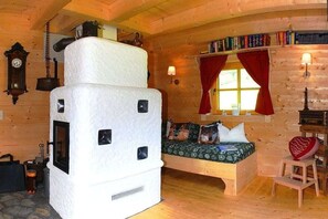Ein gemütliches Blockhaus-Interieur mit einem großen weißen Herd, einem hölzernen Bett mit gemusterten Betttextilien, einem Bücherregal und einer Wanduhr.