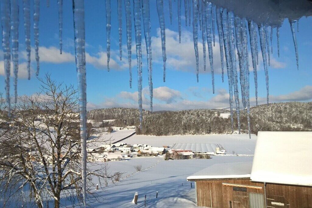 A snowy landscape with icicles hanging from a roof, a clear sky, and a distant view of a snow-covered village.