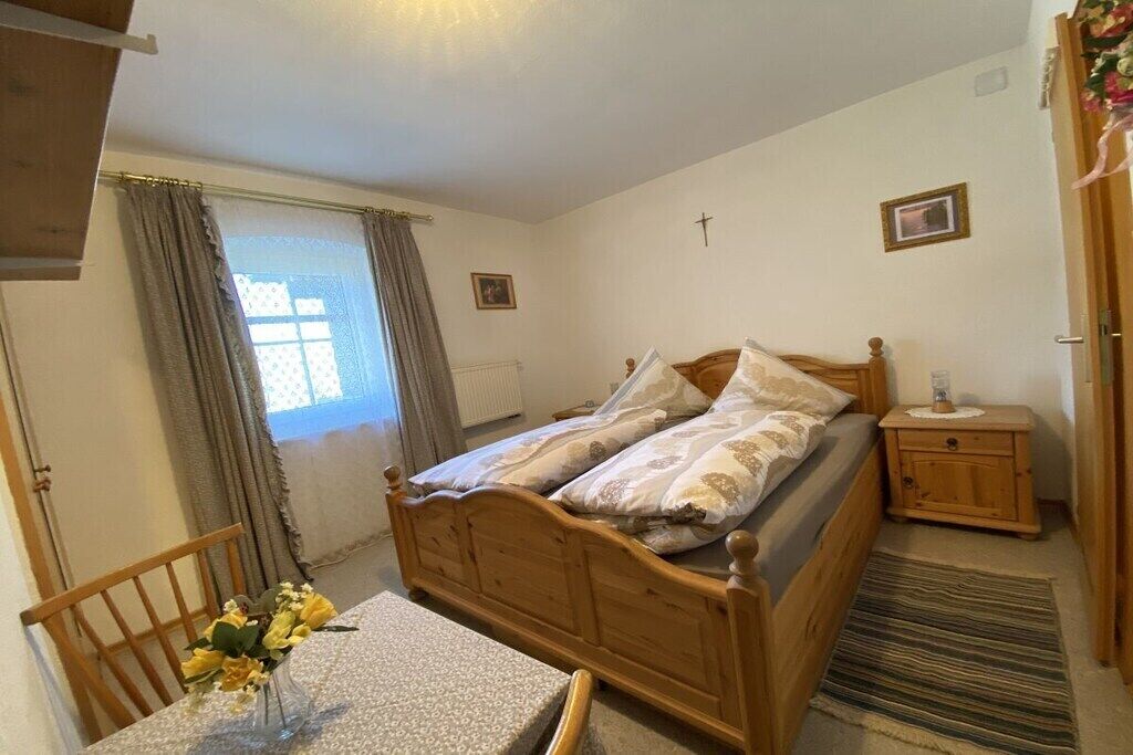 Quarto com cama de madeira, mesa de cabeceira, cadeira e janela com cortinas.