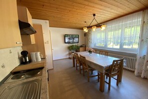 Eine Küche mit Holzbalkendecke, einem Esstisch mit Stühlen, einem an der Wand montierten Fernseher und einem Fenster mit Vorhängen.