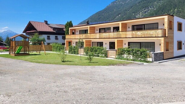 Ein zweistöckiges Gebäude mit einem Holzbalkon, einem Spielbereich für Kinder inklusive Rutsche und einem Berg im Hintergrund.