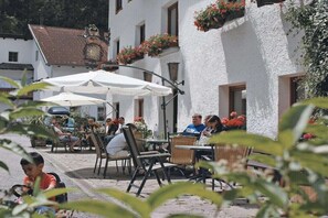 Een terras met tafels en stoelen, omgeven door witte gebouwen met bloembakken.