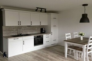 A modern kitchen with white cabinetry, a built-in oven, and a dining area with a wooden table and white chairs. - Holiday Village 39 B (Boltenhagen)