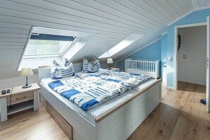 A bedroom with a sloped ceiling, a bed with a blue and white patterned bedspread, a nightstand with a lamp, and a window with a view of greenery. - Holiday Village 39 B (Boltenhagen)