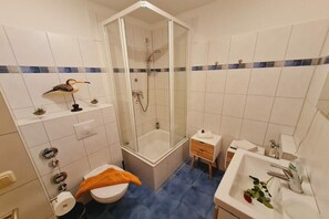 Baño con ducha envuelta en vidrio, inodoro, lavabo y una mesita de noche pequeña.