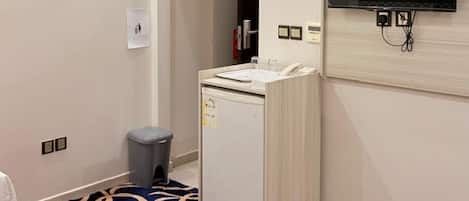 A hotel room with a bed, a television mounted on the wall, a small refrigerator, and a waste bin.