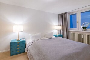 Ein Schlafzimmer mit einem Bett, zwei Nachttischen mit Lampen, einem Fenster mit Vorhängen und einer Heizung.