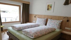 Cama doble con ropa de cama floral, una ventana con cortinas, una mesita y una lámpara de pared.