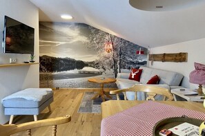 A cozy room with a large mural of a snowy landscape, a flat-screen TV, a wooden coffee table, a sofa with cushions, and a small round table. - Holiday apartment with 2 bedrooms (Füssen)
