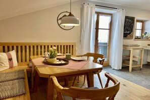 Een gezellig eetgedeelte met een houten tafel, stoelen en een bank. Boven de tafel hangt een lamp en er is een raam met gordijnen.