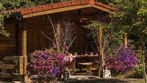 Eine rustikale Holzhütte mit einer überdachten Veranda, einem Tisch mit Stühlen und leuchtend violetten Blumen in Pflanzgefäßen.