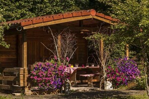 Een rustieke houten cottage met een overdekte veranda, een tafel met stoelen en levendige paarse bloemen in potten.