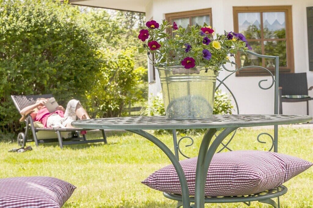 A metal table with a glass vase of flowers, a chair with a checkered cushion, and a person reclining in the background.