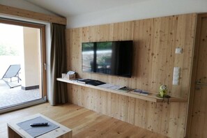 Une chambre moderne avec un mur en bois, un téléviseur fixé au mur, une petite étagère avec des livres et une figurine, et une porte-fenêtre coulissante donnant sur l’extérieur.