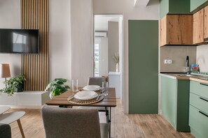 A modern kitchen with green cabinets, a wooden dining table set for two, and a flat-screen TV mounted on the wall. - Riviera City Scape 1BR & Balcony. Athens (Glifada)