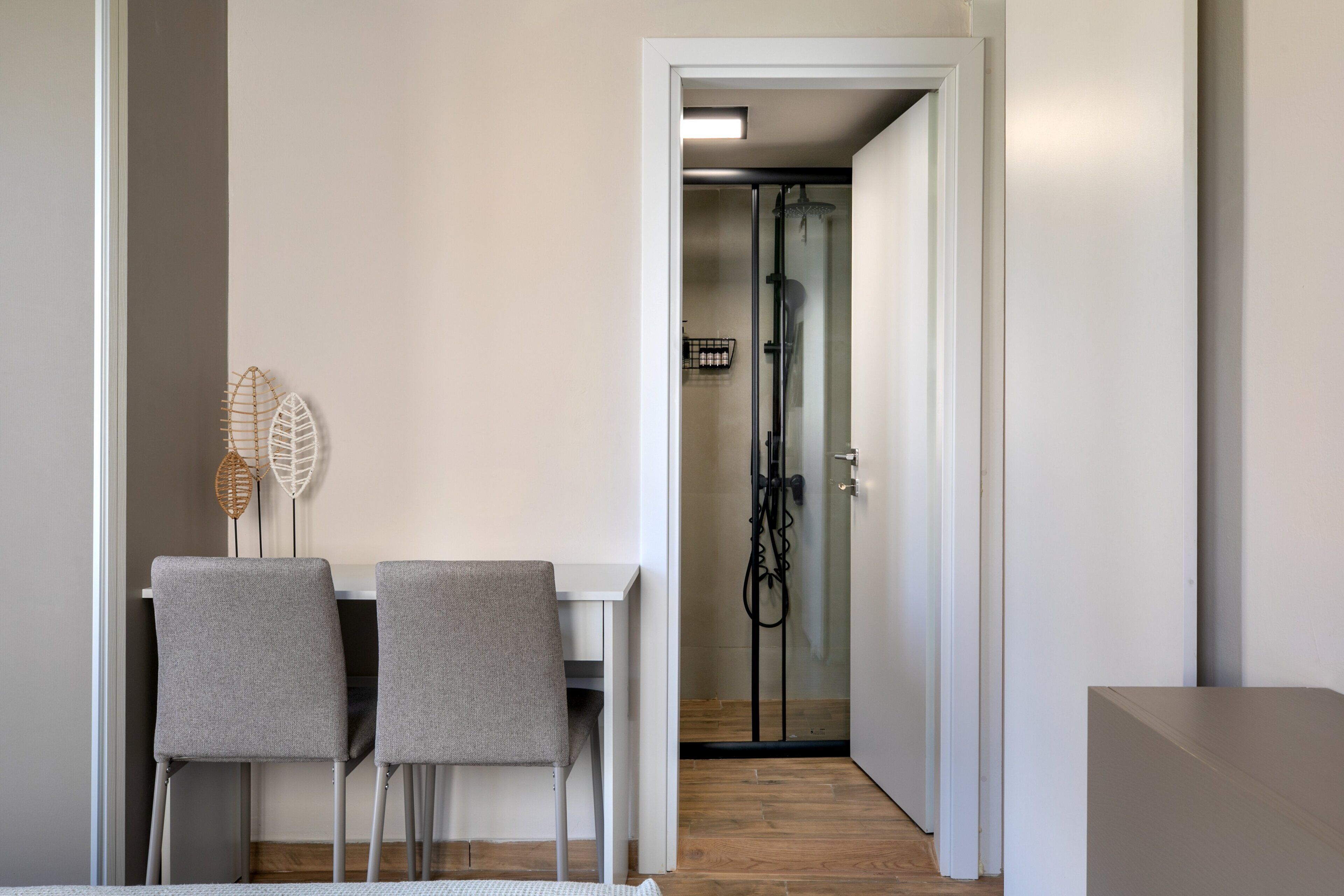 A modern interior with a dining area featuring two grey chairs and a white table, a hallway leading to a shower area, and a wooden floor.