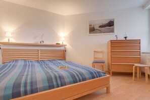 Een slaapkamer met een houten bed, nachtlampjes, een stoel, een dressoir en een ingelijst schilderij aan de muur.