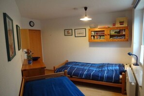 Ein kleines Zimmer mit zwei Einzelbetten, einem Holzschrank mit Büchern, einer Uhr und einer Heizung.