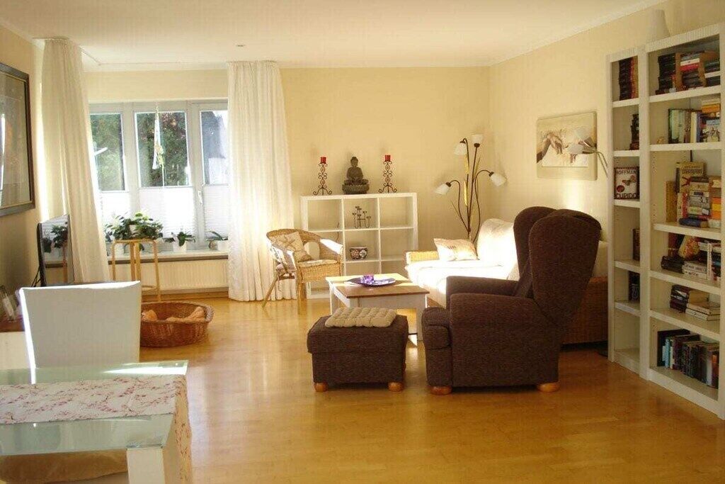 A living room with a sofa, armchair, and ottoman, a bookshelf, a white cabinet, and a window with curtains.