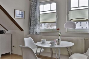 Una habitación con una mesa redonda, sillas blancas, un radiador y una ventana con vista a edificios.