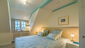 Ein Schlafzimmer mit schrägverdachter Decke, einem großen Bett mit passenden Kissen, einem Nachttisch mit Lampe und einem Fenster mit Vorhängen.