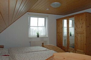 A bedroom with a wooden ceiling, a bed with a floral bedspread, a wooden wardrobe, and a window with a plant. - Holiday home with 2 bedrooms (Wangerland-Hooksiel)