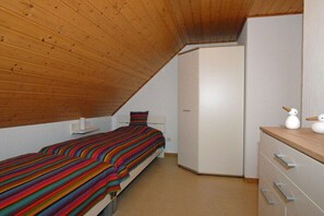 Quarto com teto inclinado, uma cama com colcha listrada e colorida, um guarda-roupa branco e uma cômoda pequena branca com objetos decorativos.