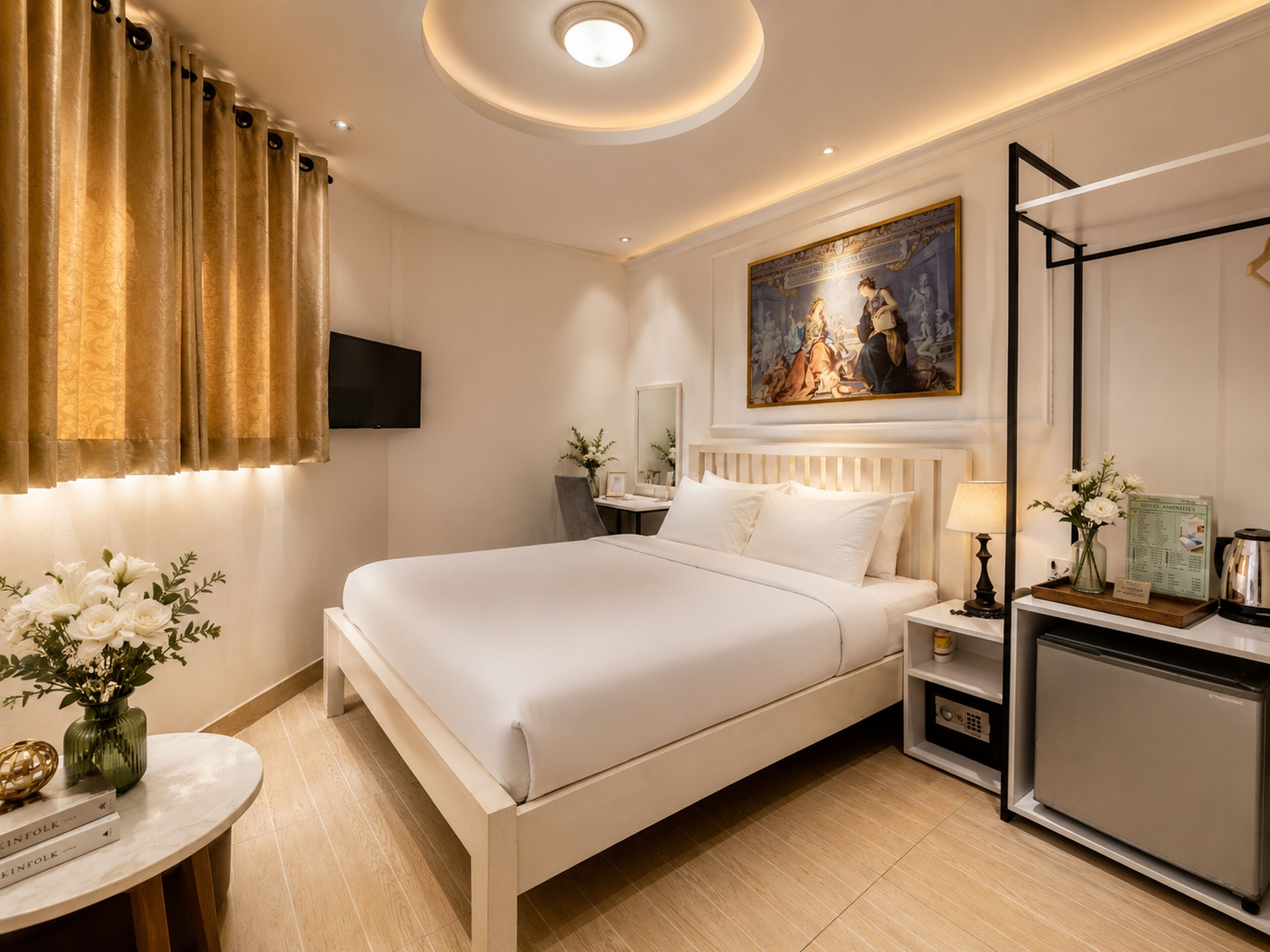 A hotel room with a large bed, a flat-screen TV, a painting above the bed, a small table with a vase of flowers, and a minibar.