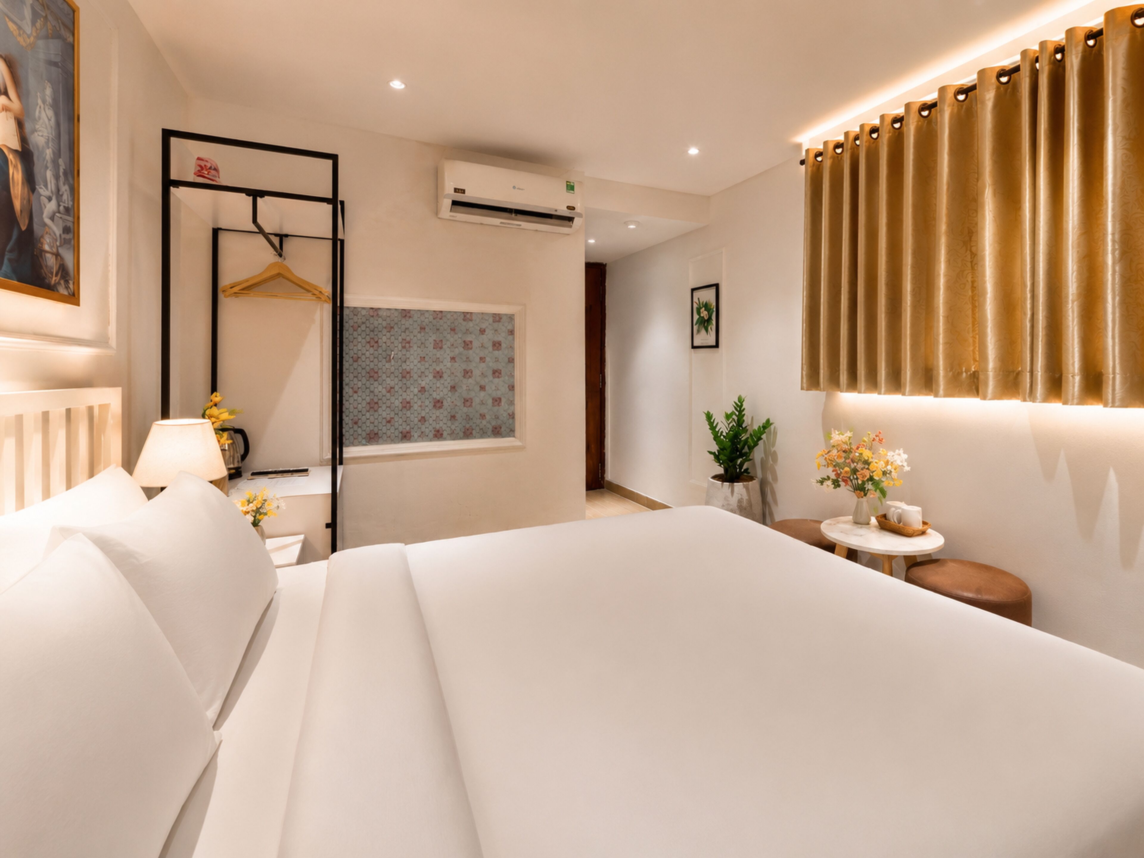 A hotel room with a bed, bedside table, lamp, air conditioning unit, window with patterned curtains, and a wall-mounted mirror.