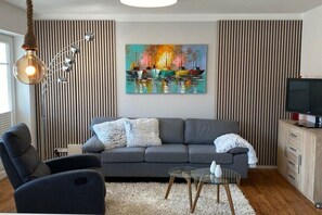 A modern living room with a grey sofa, a glass coffee table, and a painting of boats on the wall. - Holiday apartment with 1 bedroom (Bansin)