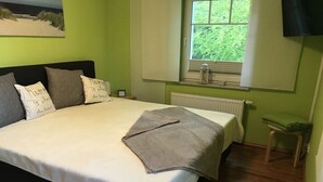 Quarto com cama, almofadas, janela com cortinas, radiador e uma pequena mesa.