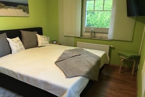 Een slaapkamer met een bed, kussens, een raam met gordijnen, een radiator en een klein tafeltje.