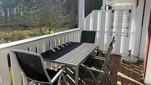 Varanda com mesa e cadeiras, corrimão branco e vista para árvores.