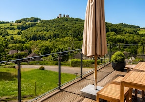 Eine Holzterrasse mit Tisch und Stühlen, die auf eine grüne Landschaft mit einem Hügel und einer Burg in der Ferne blickt.
