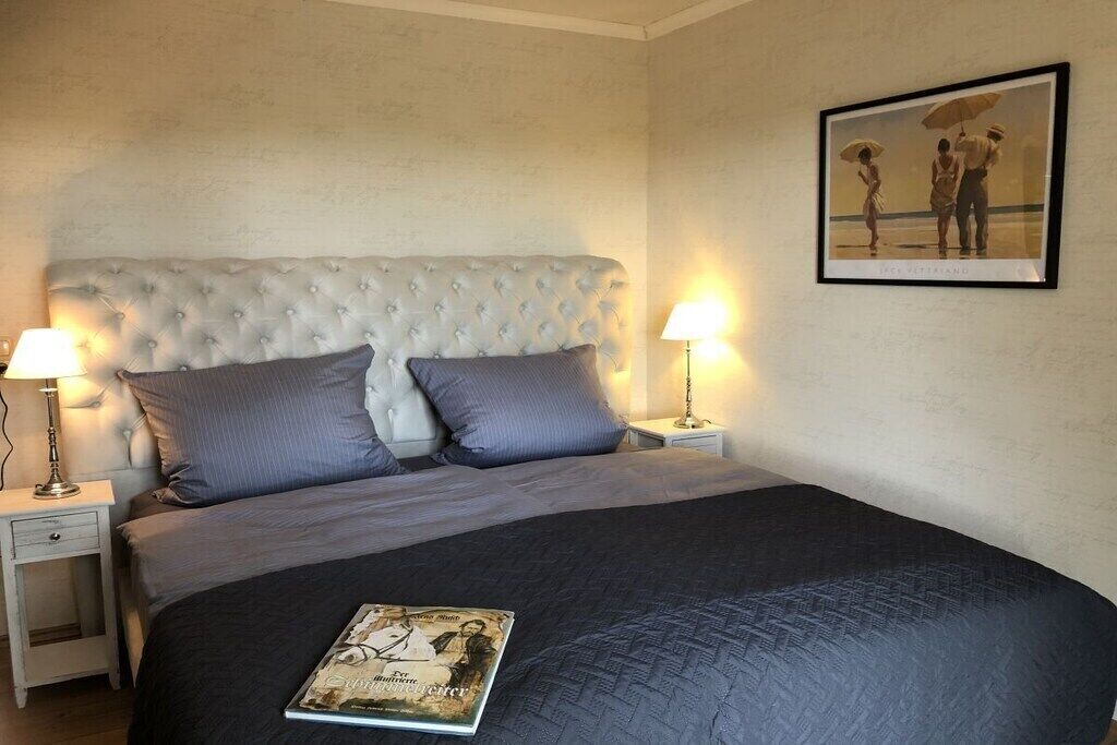 Une chambre à coucher avec une tête de lit capitonnée, deux tables de chevet avec des lampes, une couvre-lit sombre et un tableau encadré au mur.