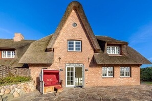 Een traditioneel bakstenen huis met een steil rieten dak, een centrale deur met glaspanelen en een stenen muur aan de linkerkant.