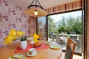 Een eetruimte met een gedekte tafel voor twee personen, een vaas met gele bloemen en uitzicht op een terras met stoelen en een tafel.