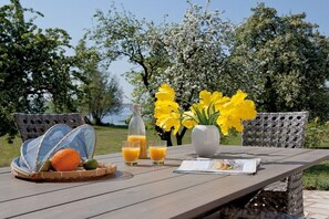 Een houten tafel met een rieten stoel, een mand met fruit, een fles, glazen jus d'orange en een vaas met gele bloemen.
